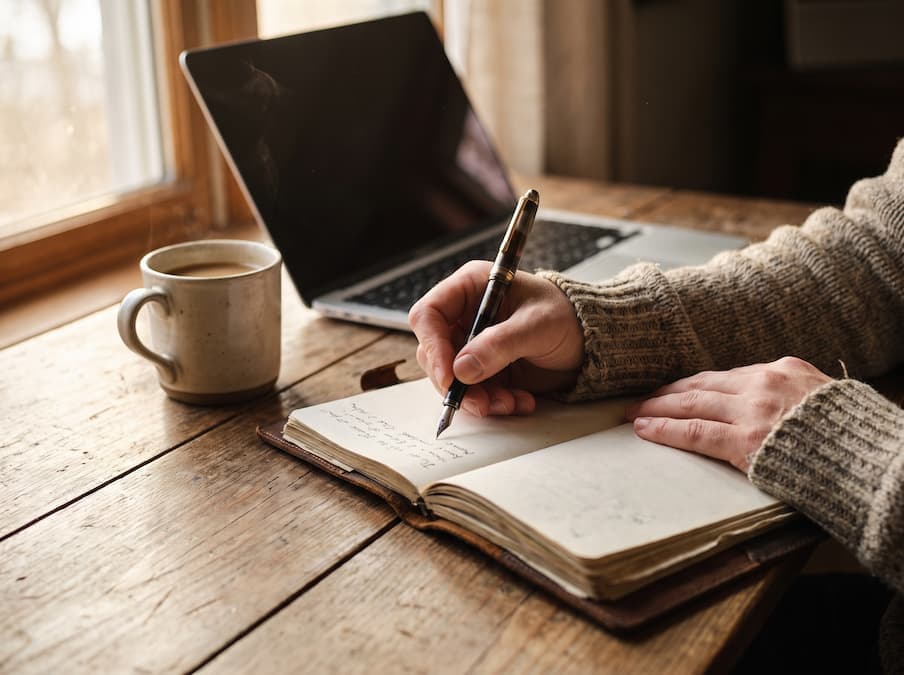 Manos escribiendo con pluma en una libreta junto a un café y un portátil, sobre una mesa de madera